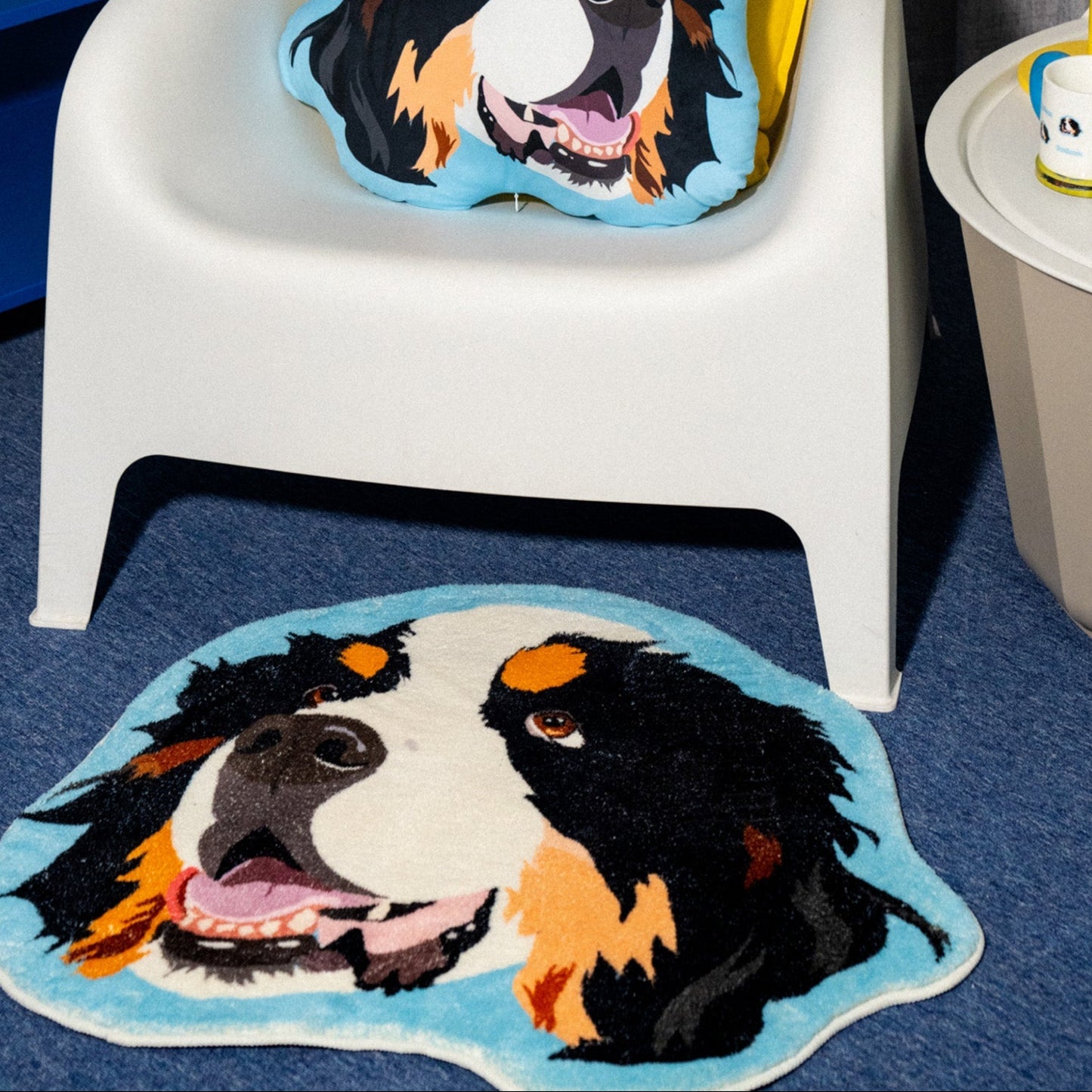 White chair with a Bernese Mountain Dog cushion, matching rug on blue carpet, and yellow lamp in the background.