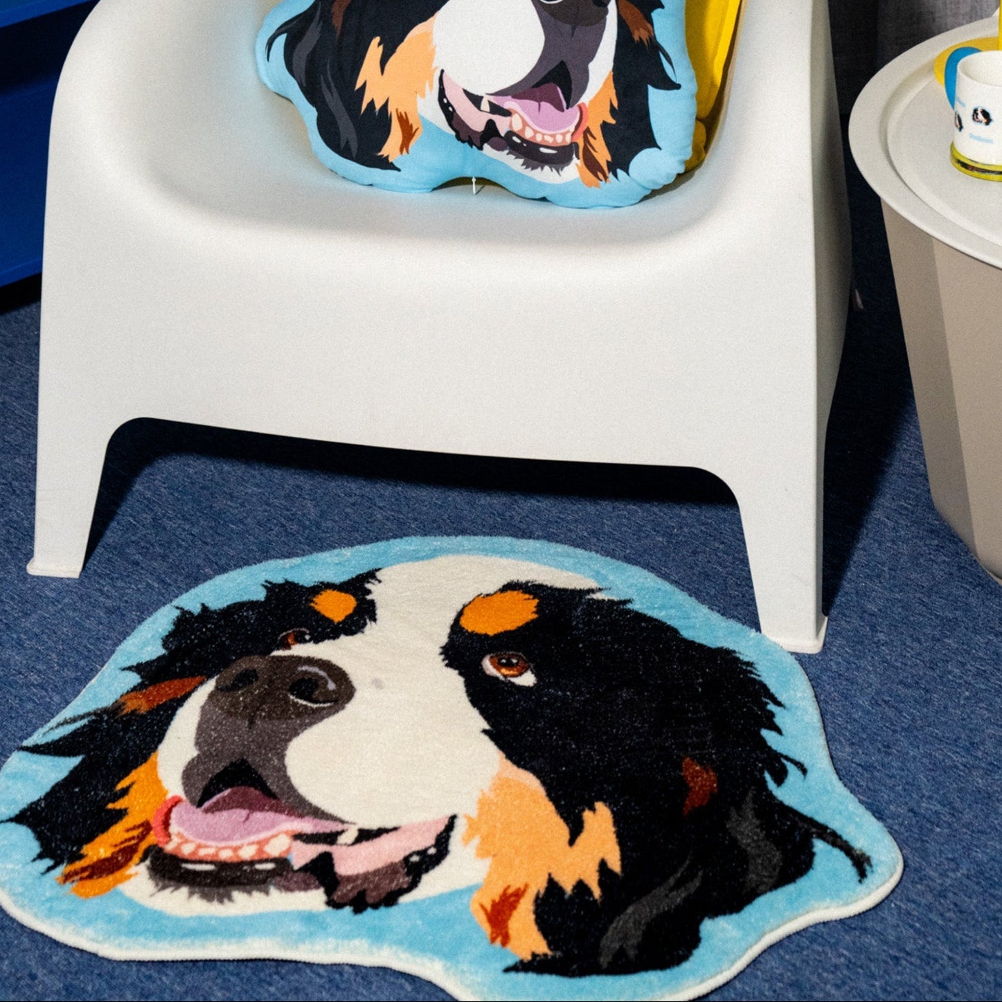 White chair with a Bernese Mountain Dog cushion, matching rug on blue carpet, and yellow lamp in the background.